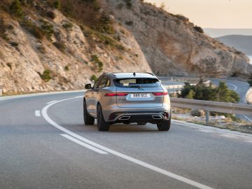 Een zilveren Jaguar F-Pace die tijdens zonsondergang over een bochtige weg naast een klif rijdt.
