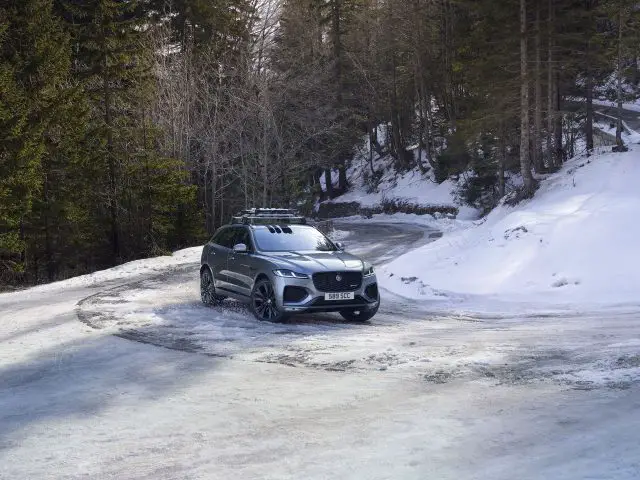 Een zilveren Jaguar F-Pace geparkeerd op een besneeuwde bosweg terwijl de zon door de bomen schijnt.