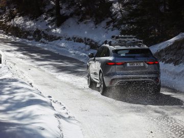 Jaguar F-Pace rijdt op een besneeuwde bergweg en spuit sneeuw opzij, onder een heldere zonverlichte hemel.