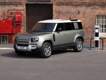 De grijze landrover Defender P400e is aangesloten op een laadstation voor elektrische voertuigen buiten een bakstenen gebouw.