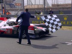 Een man in pak zwaait met een geblokte vlag naar een Toyota-raceauto die de finishlijn van Le Mans overschrijdt, met 'Rolex'-sponsorbanners op de achtergrond.
