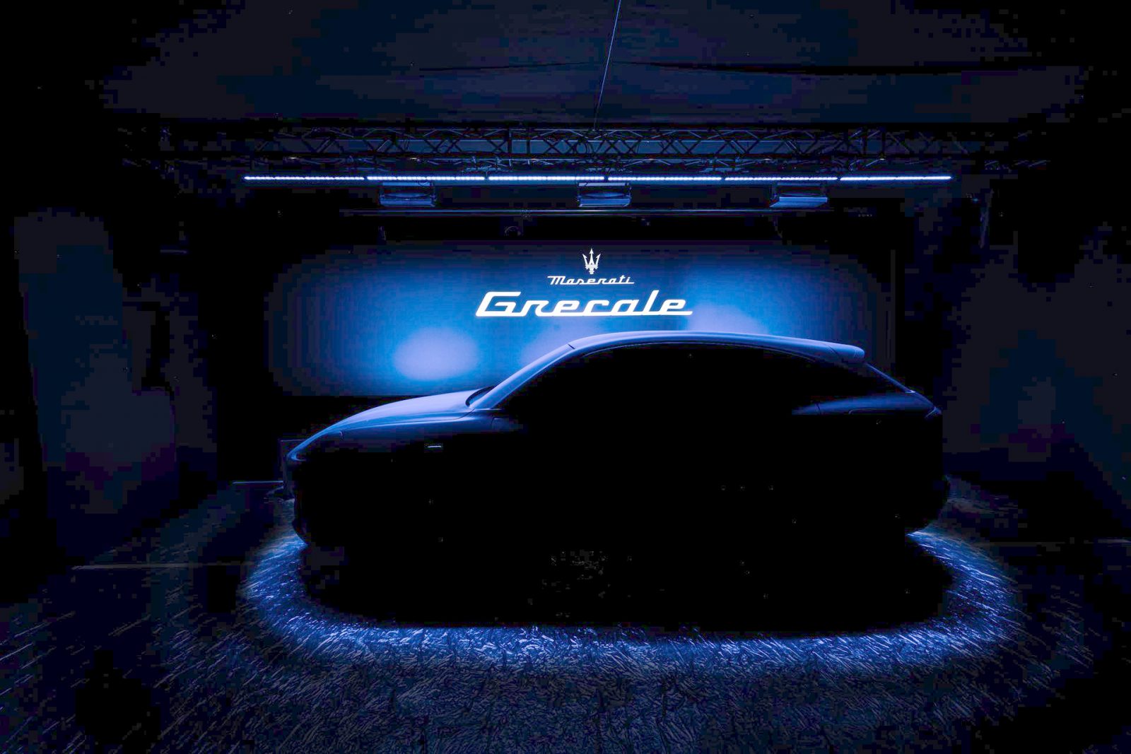 Een auto onthuld onder een spotlight met het logo "Maserati Grecale" geprojecteerd op de muur erachter, in een zacht verlichte kamer.