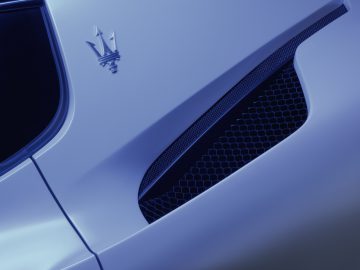Close-up van de ventilatieopening en het embleem aan de zijkant van een blauwe Maserati MC20, met de nadruk op strakke designelementen en metallic afwerking.