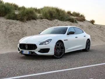 Een witte Maserati Quattroporte geparkeerd op een weg naast zandduinen.