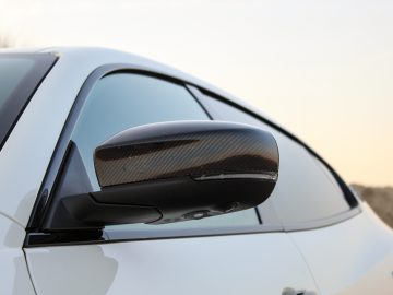 Close-up van de zijspiegel van een Maserati Quattroporte met een kap van koolstofvezel op een wit voertuig, tegen een onscherpe natuurlijke achtergrond.