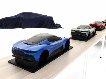 Rij schaalmodelauto's tentoongesteld op een platform, met verschillende ontwerpen en kleuren, met op de achtergrond een Maserati Grecale bedekt met een blauw zeildoek.