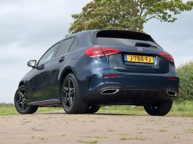 Een blauwe Mercedes-Benz A 250 e geparkeerd op een weg met een grasveld en bomen op de achtergrond op een bewolkte dag.