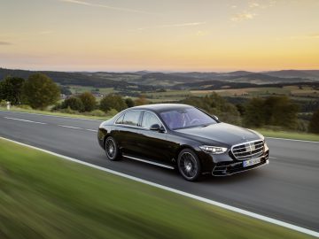 Een zwarte Mercedes-Benz S-Klasse luxe sedan die bij zonsondergang over een plattelandsweg rijdt, met glooiende heuvels en een gouden lucht op de achtergrond.