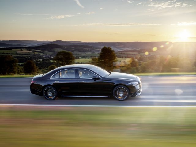 Een zwarte Mercedes-Benz S-Klasse rijdend op een snelweg met een landelijk landschap en zonsondergang op de achtergrond.