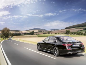 Een zwarte Mercedes-Benz S-Klasse die over een bochtige weg rijdt door een schilderachtig landschap met heuvels en een heldere hemel.