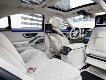 Binnenaanzicht van een moderne Mercedes-Benz S-Klasse met witleren stoelen, meerdere schermen en blauwe sfeerverlichting.