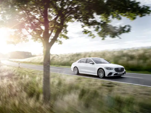 Een witte Mercedes-Benz S-Klasse sedan die bij zonsondergang op een landelijke weg rijdt met bewegingsonscherpte, wat hoge snelheid suggereert, naast een rij bomen en een veld.
