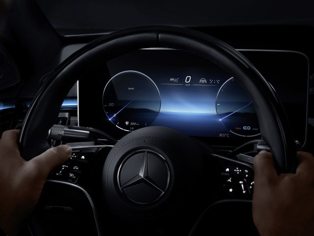Close-up van een rijdende persoon, de handen op het stuur van een Mercedes-Benz S-Klasse, met de focus op het verlichte dashboard dat de snelheid en andere voertuiggegevens weergeeft.
