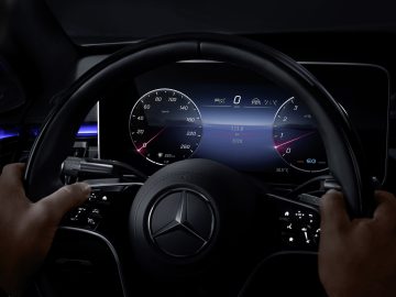 Handen van de bestuurder op het stuur van een Mercedes-Benz S-Klasse, met uitzicht op een digitaal dashboard dat de snelheid en andere voertuiggegevens weergeeft.