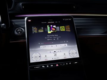 Binnenaanzicht van een Mercedes-Benz S-Klasse met een groot touchscreen-display op het dashboard, met zichtbare mediabediening en temperatuurinstellingen.