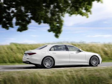 Een witte Mercedes-Benz S-Klasse luxe sedan rijdend op een weg met groene bomen en een blauwe lucht op de achtergrond.