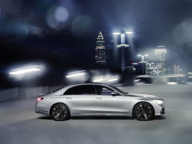 Een zilveren Mercedes-Benz S-Klasse snelt door een nachtelijk stadsbeeld met vage lichten en wolkenkrabbers op de achtergrond.