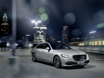 Een zilveren Mercedes-Benz S-Klasse rijdt 's nachts door een stadsstraat, met stadslichten en wolkenkrabbers verlicht op de achtergrond.