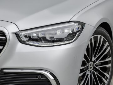 Close-up van de koplamp en het stuur van een witte Mercedes-Benz S-Klasse, met modern design en gedetailleerde styling.