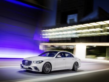 Witte Mercedes-Benz S-Klasse sedan die 's nachts rijdt met een wazige stadsachtergrond, waarbij beweging en snelheid worden benadrukt.