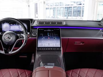 Interieur van een Mercedes-Benz S-Klasse met een modern dashboard met digitaal display, stuur en roodleren stoelen.