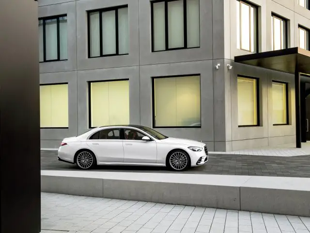 Witte Mercedes-Benz S-Klasse geparkeerd nabij een modern gebouw met grote verlichte ramen.