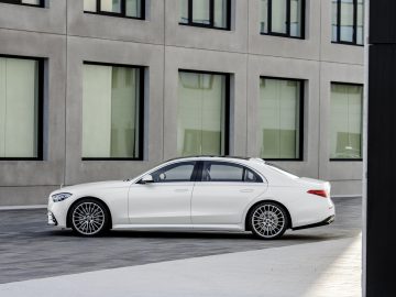 Een witte Mercedes-Benz S-Klasse geparkeerd in een modern stedelijk gebied met betonnen gebouwen en grote ramen.
