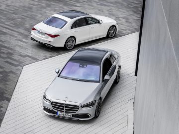 Twee Mercedes-Benz S-Klasse-auto's geparkeerd op een trottoir met patronen, van bovenaf gezien, waarvan de ene in zilver en de andere in wit is.