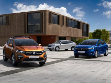 Drie Dacia Sandero-auto's in verschillende kleuren geparkeerd voor een eigentijds gebouw op een zonnige dag.