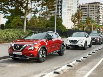 Drie Nissan Juke SUV's rijden in een stad, met de rode lijn gevolgd door een witte en een grijze SUV, tegen een stedelijke achtergrond met bomen en hoge gebouwen.