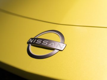 Close-up van een Nissan Z Proto-logo op de motorkap van een felgele auto, waarbij de chromen afwerking van het embleem wordt benadrukt.