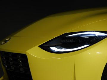 Close-up van de koplamp van een gele Nissan Z Proto en een deel van de grille op een donkere achtergrond.