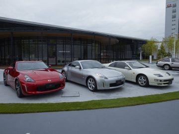 Drie Nissan-sportwagens, gaande van historisch tot de Nissan Z Proto, geparkeerd buiten een Nissan-bedrijfsgebouw onder bewolkte hemel.