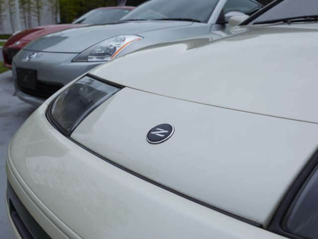 Close-up van het logo van een witte Nissan Z Proto-auto op de motorkap met andere auto's wazig op de achtergrond.