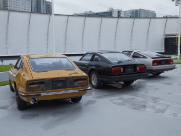 Drie klassieke Nissan Z Proto-auto's geparkeerd op een natte ondergrond, met de dichtstbijzijnde auto in een goudgele kleur.