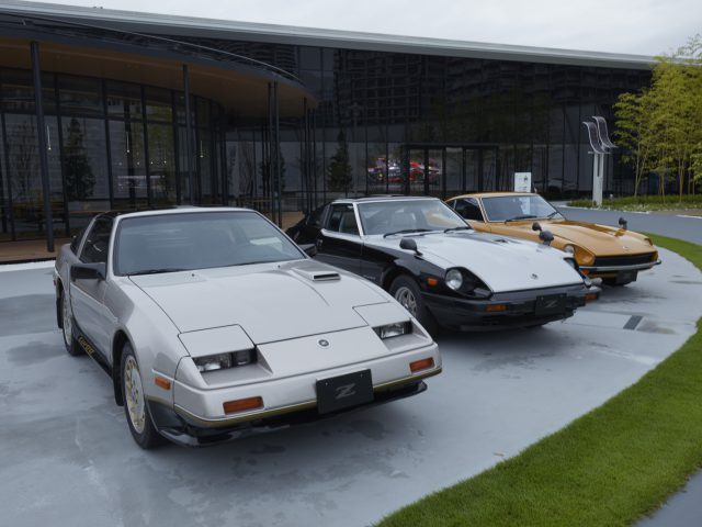 Twee vintage auto's uit de Nissan Z-serie, een witte en een gele, geparkeerd voor een modern gebouw op een bewolkte dag, die doet denken aan het Nissan Z Proto-ontwerp.