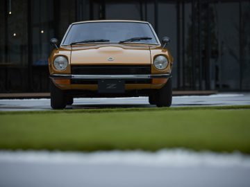 Vooraanzicht van een oranje Nissan Z Proto-auto geparkeerd op een stoep met moderne glazen gebouwen op de achtergrond.