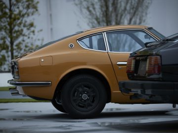Zijaanzicht van een vintage goudbruine Nissan Z Proto geparkeerd op een betonnen stoep, met de achterkant van een andere auto zichtbaar op de voorgrond.