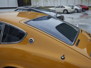 Close-up van de achterruit en vin van een gele Nissan Z Proto, met wazige auto's op de achtergrond op een parkeerplaats.