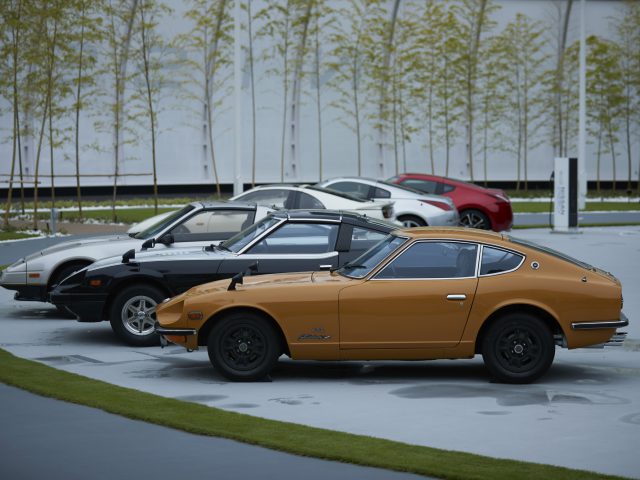 Een rij vintage sportwagens, waaronder een goud-zwarte Nissan Z Proto, geparkeerd op een modern terrein met netjes gerangschikte bomen op de achtergrond.