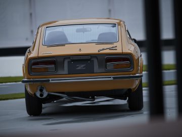 Achteraanzicht van een vintage oranje Nissan Z Proto-auto met chromen bumpers geparkeerd voor een wit gebouw.