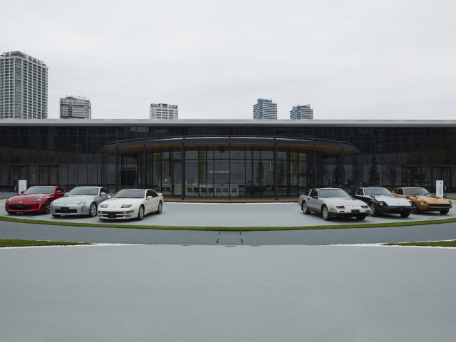 Vijf Nissan Z Proto-sportwagens geparkeerd voor een modern rond gebouw met een stedelijke skyline op de achtergrond.