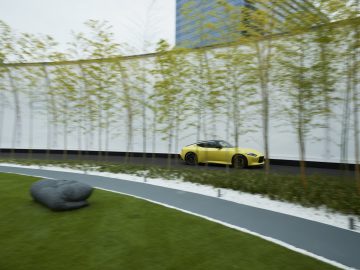 Een gele Nissan Z Proto-sportwagen geparkeerd op een smalle weg, te midden van een moderne tuin met hoge, dunne bomen en hoge gebouwen op de achtergrond.