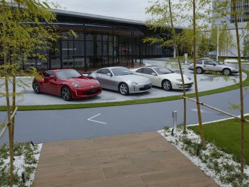 Luxe auto's, waaronder de Nissan Z Proto, geparkeerd op een keurig geplaveid terrein buiten een modern glazen gebouw, omgeven door jonge bomen en aangelegde tuinen.