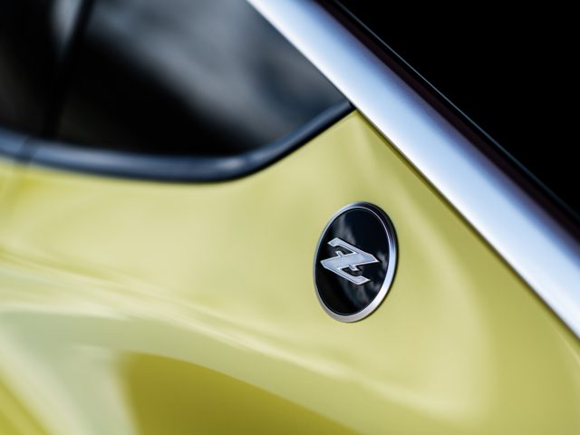Close-up van een gele Nissan Z Proto met een zwart en zilver 'z'-embleem op de carrosserie.