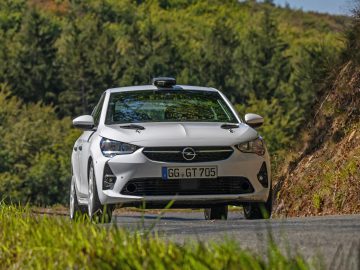 Witte Opel Corsa Rally4 rijdt op een gebogen weg met dichte groene bomen op de achtergrond op een zonnige dag.