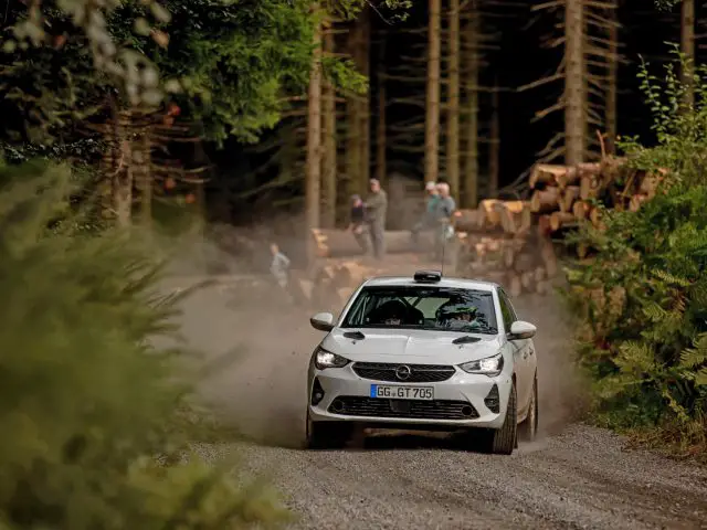 Een Opel Corsa Rally4 rijdt over een stoffige bosweg terwijl omstanders vanaf de zijkanten toekijken bij gestapelde boomstammen.