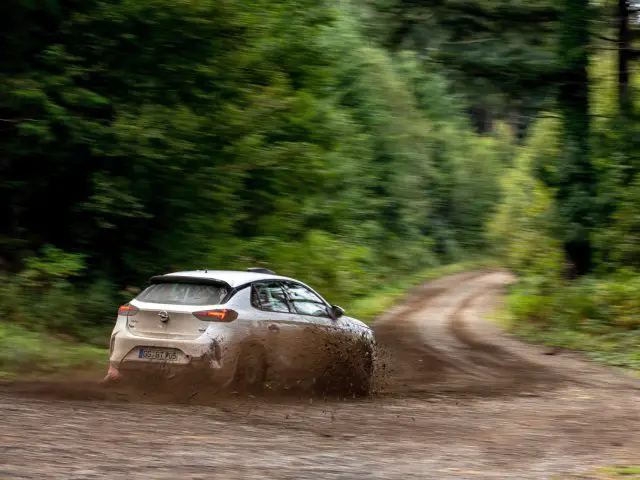 Een Opel Corsa Rally4 snelt over een modderige bosweg en stuwt water en modder uit een plas, in een wazig, bewegingsrijk tafereel.