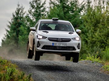 Een witte Opel Corsa Rally4-auto die lucht opvangt terwijl hij over een onverharde weg in een bosrijk gebied snelt, met stof achter zich aan.