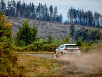 Een witte Opel Corsa Rally4 racet op een stoffige grindweg in een landelijk gebied met pijnbomen op de achtergrond en een bewolkte lucht.